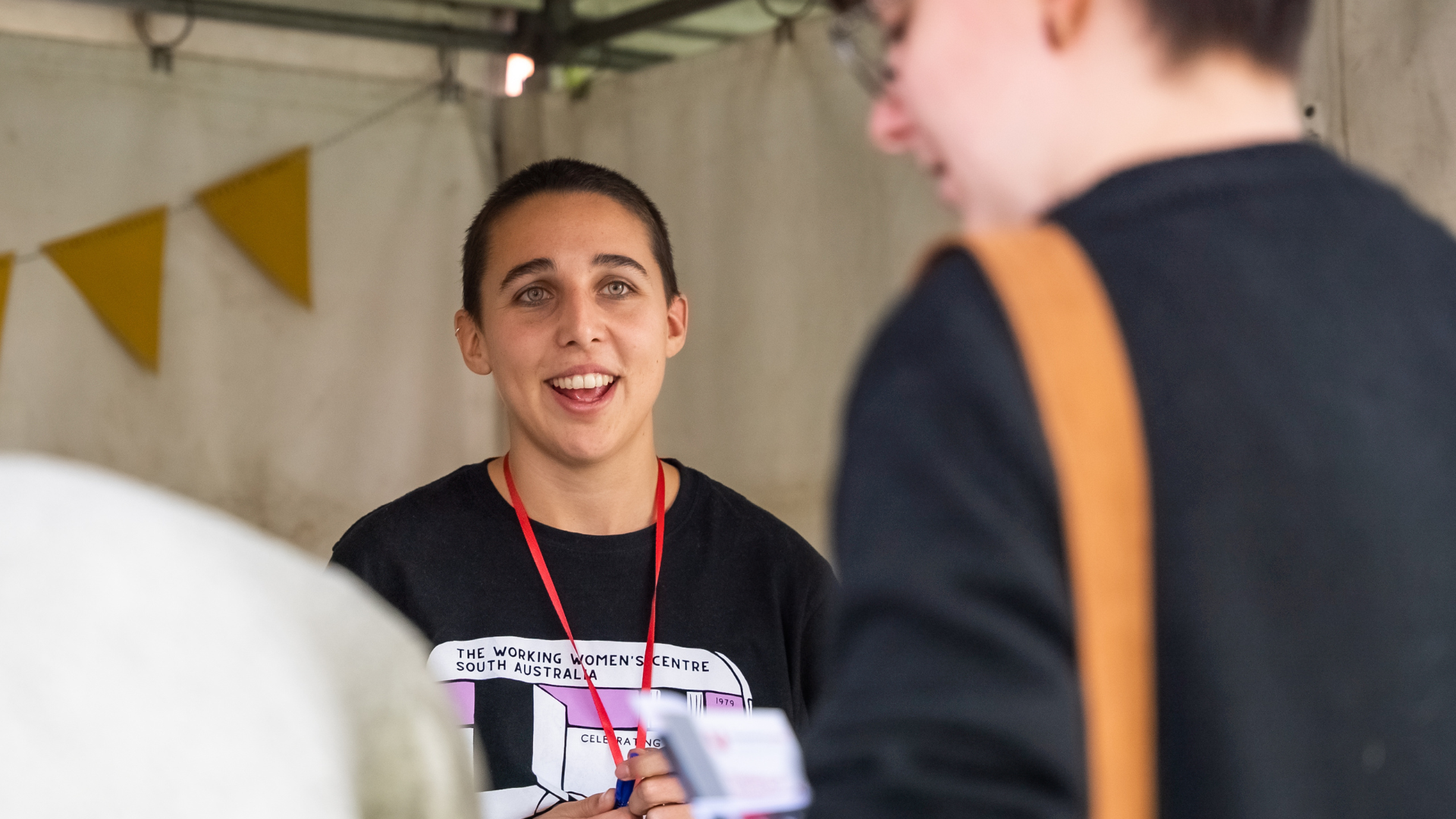 A volunteer with short hair and a black t-shirt talks with a student behing an O'Week stall